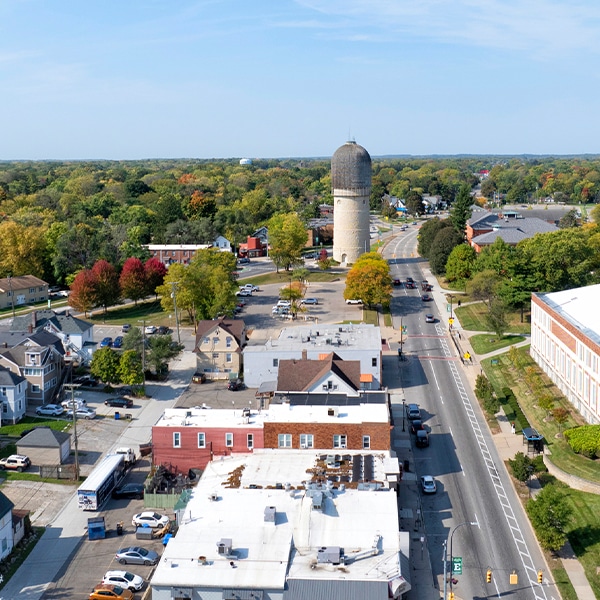 Aerial view of a small town street with a tall, round brick silo and surrounding buildings, set against a green landscape.